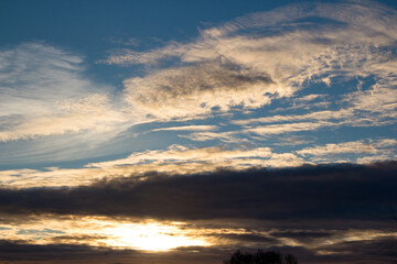 Golden hour glow peeking through dramatic dark clouds, with lighter wispy formations above, creating a serene and captivating sky scene