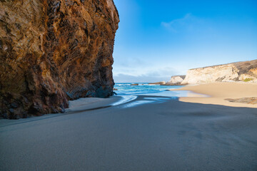 Scenic sandy beach with golden cliffs and turquoise ocean waves under bright blue sky showing contrast between smooth sand, textured rock formations, and gentle sea water reflections in sunlight