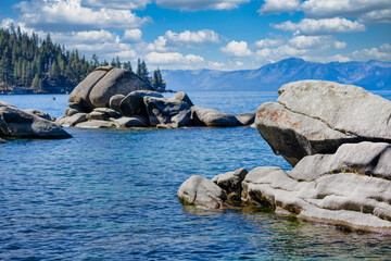 Granite boulders along the shore of Bonsai Rock at Lake Tahoe Nevada USA with clear blue water, pine trees and Sierra Nevada mountains under bright summer sky with white clouds on a calm sunny day