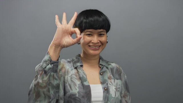 Chinese woman smiling making ok gesture in grey background showing positive emotion and confidence with isolated expression indicating approval.