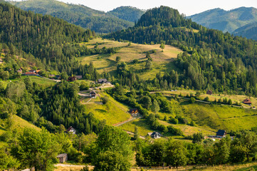 Lush green valley with rural houses surrounded by forested mountains under clear blue sky.