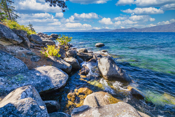Lake Tahoe California USA scenic rocky shoreline with clear blue water and large granite boulders under bright sky and white clouds summer view of pristine Sierra Nevada nature and landscape