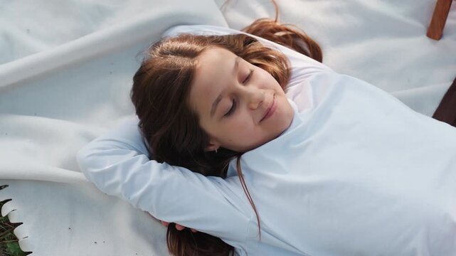 Aerial view of young girl lying faced up thoughtfully on soft white blanket outdoors, gazing into distance with calm expression, peaceful mood, sunlight highlighting her hair, evoking relaxation