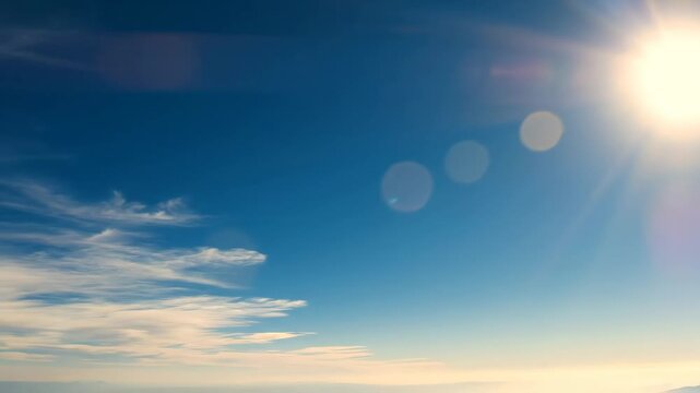 Bright Sunlight in Blue Sky with Clouds - A bright sun shines in a light blue sky with a few scattered white clouds. The sun is creating a lens flare effect with a few bokeh circles.