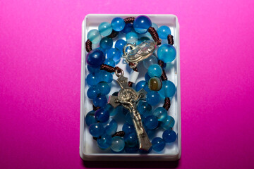 A blue rosary with twisted cords and large glossy beads rests in a small white box. The metal crucifix of Jesus Christ and the silver-toned medallion emphasize the religious purpose of the object.
