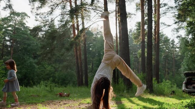 Young miss performing energetic cartwheel on picnic mat in sunny forest while little sister stands nearby watching playfully, surrounded by tall trees and warm sunlight