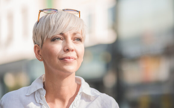 Beautiful middle aged woman with blond short hair wearing sunglasses. Profile of female person dressed in white summer shirt. Outdoors-outside