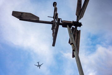 A unique contrast between old, wooden directional weather vane and a modern commercial jet airplane silhouetted against a dramatic cloudy sky. Symbolizes technology, travel, environment, and movement.