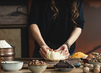 A person with long hair is kneading dough on a wooden board. Several bowls around the dough contain ingredients like chocolate, nuts, and flour. It looks like they're baking something delicious