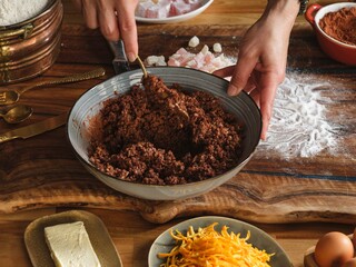 Hands mix a brown batter in a bowl, combining it with ingredients on a table. Flour, Turkish delight, shredded orange peel, and butter are spread around for baking