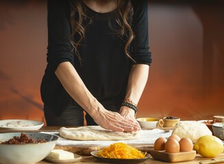 A woman in a black shirt prepares a meal. She flattens dough on a wooden board surrounded by ingredients like cheese, eggs, lemon, and a bowl of savory filling in warm light