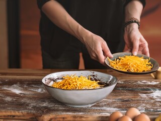 A person is adding shredded carrots from a small plate into a bowl. This preparation is for making a sweet treat in anticipation of the Thanksgiving holiday. Eggs are near