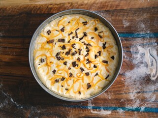 A delectable cheesecake sits in a metal pan. Orange peel zest dances with dark chocolate pieces, all on top of a creamy, baked filling. Flour dusts the wooden surface