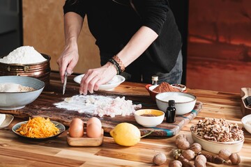 In a warm kitchen, a person chops candied fruit on a wooden board surrounded by bowls of flour, eggs, nuts, lemon, and spices, preparing to bake holiday treats