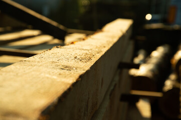 Bright sunlight illuminates the rough surface of a wooden plank at a lumber yard, highlighting the natural texture and warmth of the wood