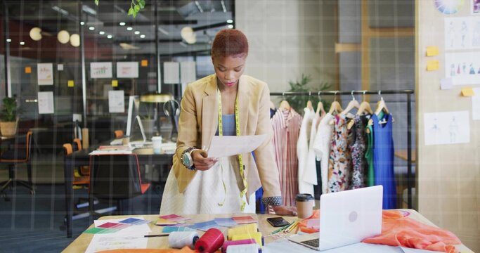 Fashion designer in blazer and dress reviewing document in studio, with fabric swatches and laptop