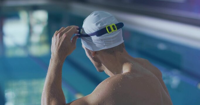 Adult male swimmer adjusting swim cap while holding goggles in aquatic facility with marked lanes