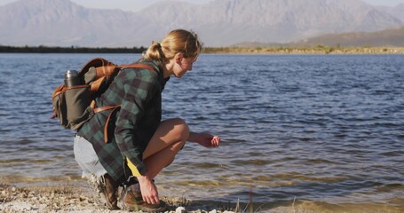 Crouching hiker touching lake water on pebbled shore with backpack, boots, water bottle, copy space