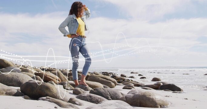 Shading eyes, woman wearing denim jacket cuffed jeans gazing across beach rocks, waves, copy space