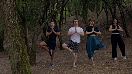 Four young friends engage in a mindful yoga session, performing the tree pose amidst the tranquility of a lush forest. This serene outdoor activity promotes balance, fitness, and overall well-being.