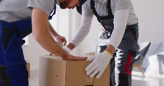 Cropped view of two professional movers sealing cardboard box with duct tape while preparing items for relocation. Male workers of moving company packing belongings during move, ensuring safe delivery