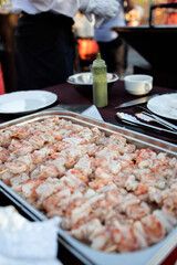 A metal tray filled with raw shrimp skewers, ready for grilling, set on a dining table. Accompanied by a green sauce bottle, cutlery, and plates. In the background — a chef and a glowing grill, creati