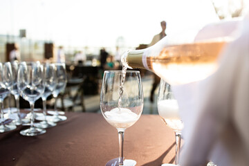 A bottle of rosé wine is being poured into a glass at an outdoor event. Several empty wine glasses are lined up on a table in the background, creating a festive atmosphere under the warm sunlight.