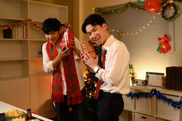 Two coworkers joyfully shake wrapped gifts, guessing what's inside during a cheerful Christmas celebration on the final day at work