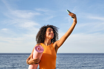 Smiling woman taking selfie with smartphone, holding yoga mat at the sea, enjoying wellness moment
