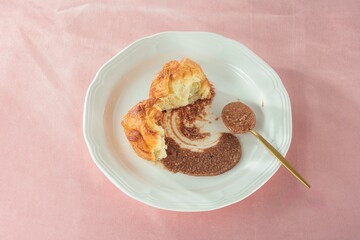 A torn golden pastry sits on a white plate with a smear of chocolate spread. A gold spoon is filled with more spread
