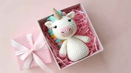 Canvas schilderij Eenhoorn A delightful crocheted unicorn toy nestled in a pink gift box with matching ribbon, creating a heartwarming scene of a handmade present on a pink background  © MdNayan