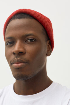 Portrait of young Black man looking into camera wearing red knit hat and shirt, showing neutral facial expression against plain background, close up framing