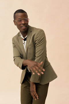 Young Black man wearing sunglasses dancing and smiling while looking away from camera, standing against plain background, showing expressive hand gesture and confident body language