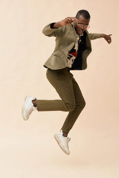 Black man jumping mid air with energetic expression, wearing sunglasses, posing against plain background, showing dynamic movement and stylish attitude