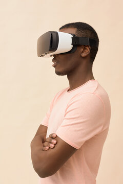 Young Black man standing with arms crossed wearing virtual reality headset, facing sideways and engaging with immersive digital experience against plain background