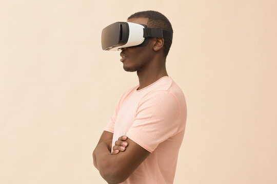 Young Black man standing with arms crossed wearing virtual reality headset, engaging with immersive digital experience against plain background, side profile visible