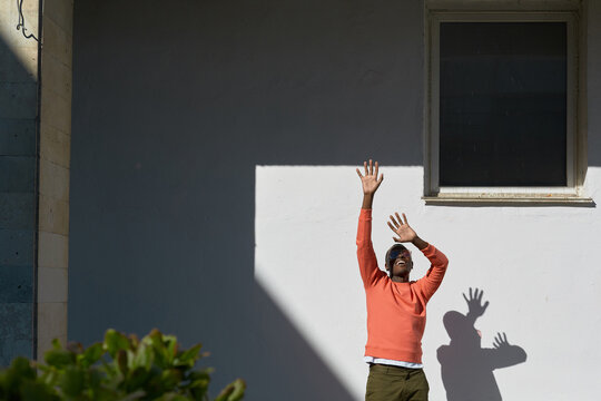 African American man  standing outdoors raising arms toward sunlight casting distinct shadow on wall background, facial expression showing excitement and engagement