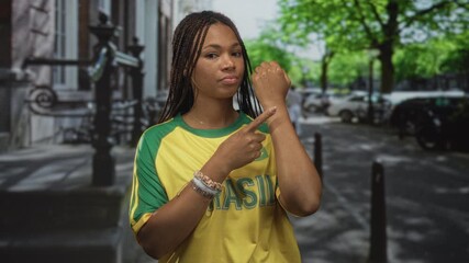Woman wearing yellow brazil jersey and bracelets points finger to wrist while standing on city street; time awareness pride. - Powered by Adobe