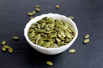  On a black background there is a white bowl with peeled pumpkin seeds.	