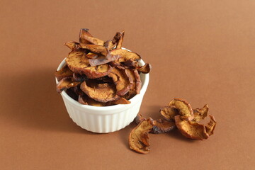 On a brown background lies a small pile of dried apple pieces in a bowl.