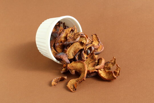 On a brown background lies a small pile of dried apple pieces in a bowl.