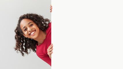 A happy African American preteen girl is smiling as she poses with a blank white board against a gray background. She looks excited and ready to showcase your message on the empty poster.