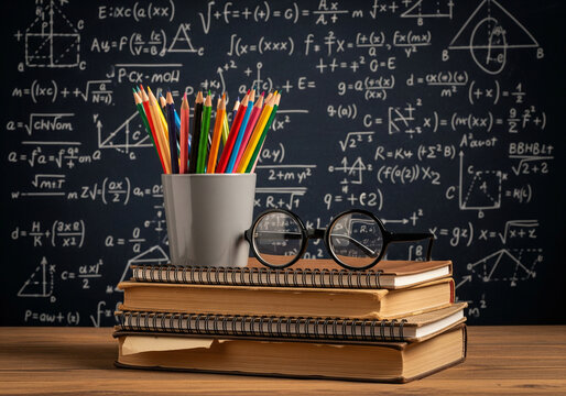 A stack of old books with colorful pencils in a holder and round glasses in front of a chalkboard covered in math equations - Powered by Adobe