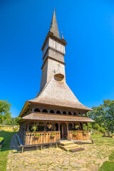 Surdesti church of Archangels Michael and Gabriel in Maramures, Romania, tall shingled spire and bell tower of a historic UNESCO World Heritage church under blue sky in Maramures, Romania