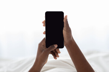 Closeup of female hands holding a modern black smartphone with a blank screen. The unrecognizable woman is relaxing in bed, using the device to access mobile applications.