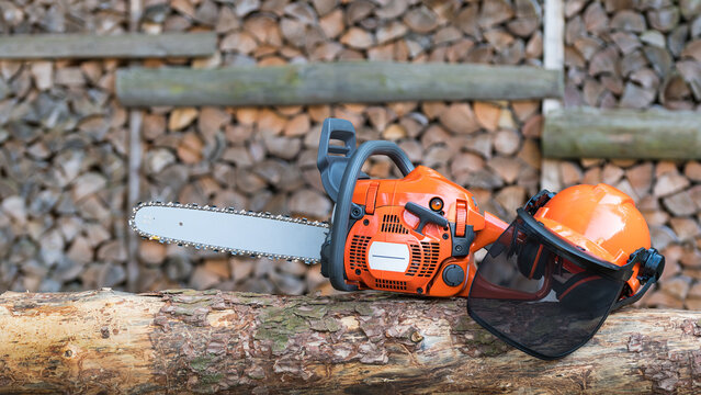 Portable gasoline chainsaw and helmet with protective mesh visor on wooden log. Professional equipment for safety cutting wood on felled tree trunk with brown green bark on blurry firewood background.