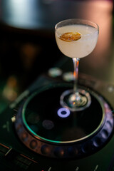 A sophisticated cocktail with a citrus slice garnish, presented in a coupe glass on a professional DJ turntable in a dimly lit club or bar.