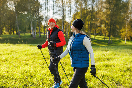 A man and a woman dressed in sporty autumn gear walk with Nordic poles across a grassy park on a bright, sunny day, enjoying fitness in nature