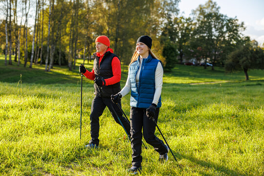A man and a woman dressed in sporty autumn gear walk with Nordic poles across a grassy park on a bright, sunny day, enjoying fitness in nature - Powered by Adobe
