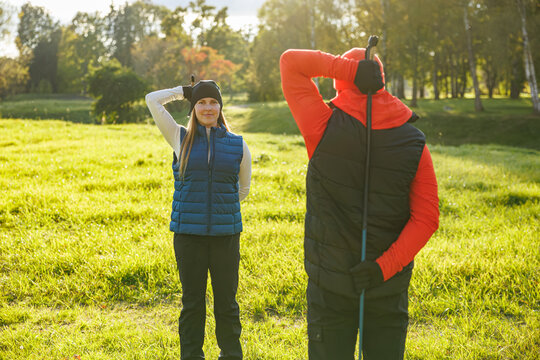 A smiling woman and a man in outdoor gear stretch in a sunny grassy field, preparing for a Nordic walking session with poles, surrounded by trees and open nature